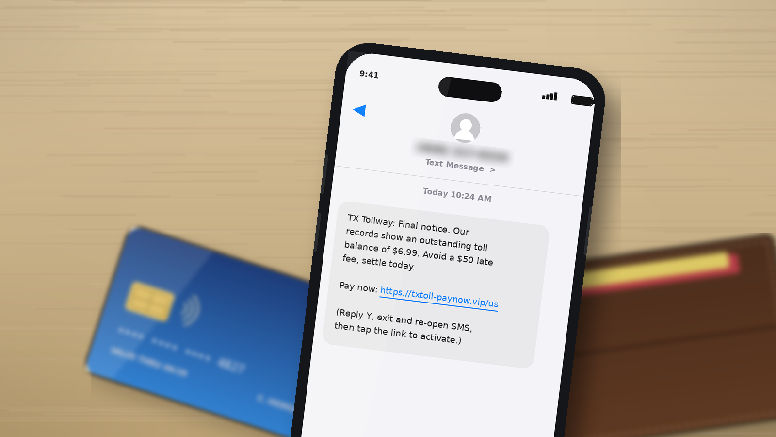 A close up of a smartphone screen showing a suspicious text message about an unpaid toll, with a blurred credit card and laptop in the background.